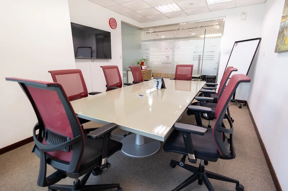 Spacious meeting room with a large conference table, red chairs, wall-mounted screen and windows